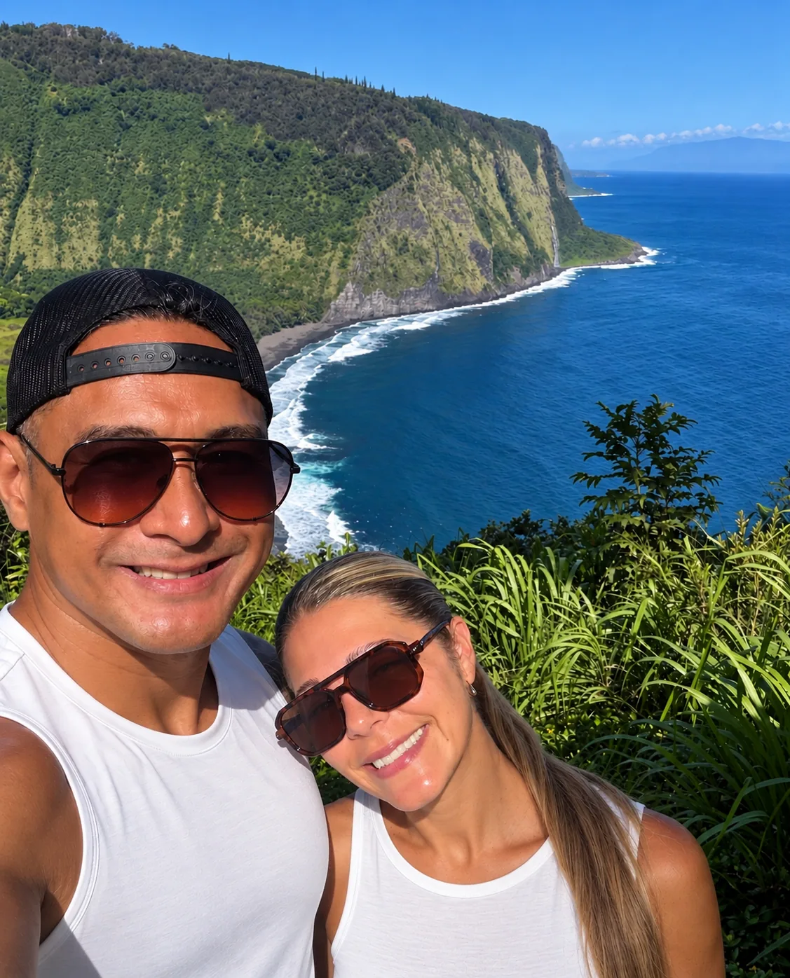 Photograph of Talanui co-founders Chris and Tiffany Paraoan smiling together at a coastal overlook, with green cliffs and the ocean behind them.